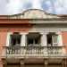 Da qui riusciamo a scorgere meglio i decori della facciata. Nella parte centrale spicca il balcone in pietra, oltre il quale intravediamo quattro bianche colonne scanalate...